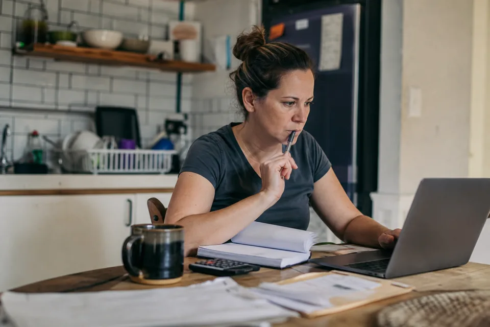 Small business owner at a kitchen table reviewing a website subscription on a laptop next to a stack of invoices