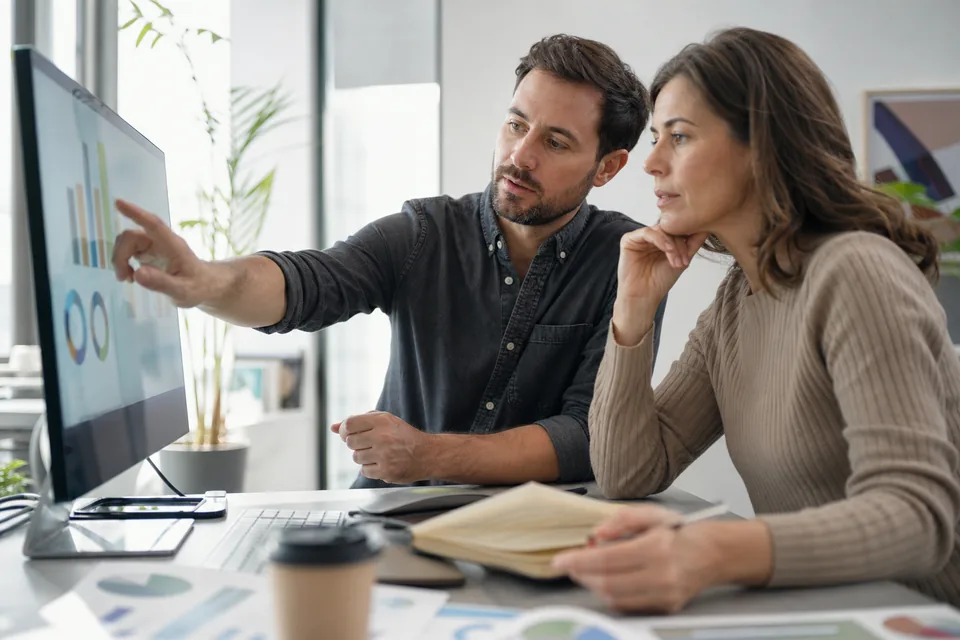 Two paid media marketers comparing landing page traffic and ad spend on a large monitor in a small office